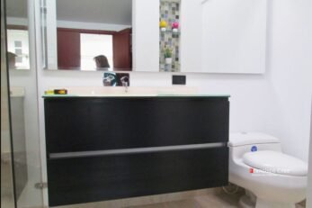 Un mueble de baño flotante de color negro satinado, fabricado a medida con tableros RH resistentes a la humedad. Cuenta con cajones de cierre lento con herrajes Blum/Häfele, un mesón de cuarzo negro y un lavamanos moderno de sobreponer. La grifería negra mate complementa el diseño.