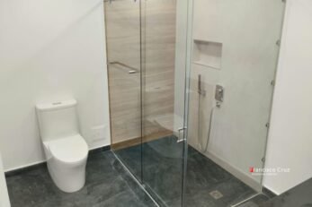 Detalle de piso y division de baño con cerámica gris de textura antideslizante en el 100% del área. Incluye una ducha a ras de piso con barra de apoyo de seguridad, diseñada para reducir el riesgo de caídas y facilitar el uso autónomo.