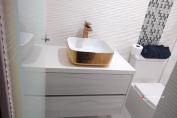 Primer plano de un mueble de baño moderno de color blanco puro, fabricado a medida con tableros RH y cajones con herrajes de cierre lento certificados. Sobre el mesón de cuarzo blanco descansa un llamativo lavamanos de sobreponer en tono poma rosa pastel, que se convierte en el punto focal del diseño. Grifería cromada y pared enchapada en porcelanato claro texturizado.