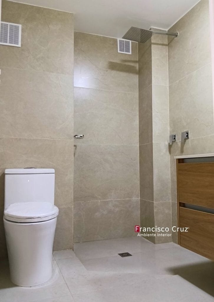 Inodoro blanco de una pieza junto a ducha con enchape beige, rejilla de piso plana y regadera cuadrada superior.