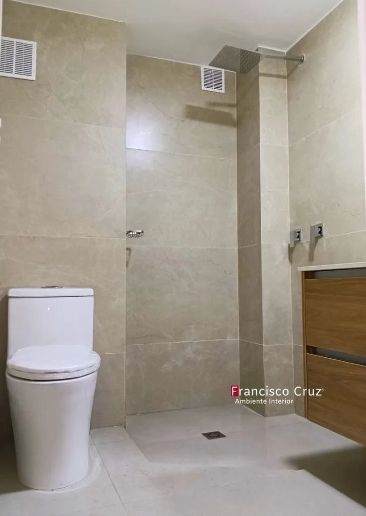 Inodoro blanco de una pieza junto a ducha con enchape beige, rejilla de piso plana y regadera cuadrada superior.